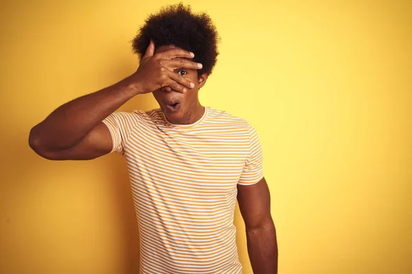 American man with afro hair wearing striped t-shirt standing over isolated yellow background peeking in shock covering face and eyes with hand, looking through fingers with embarrassed expression.