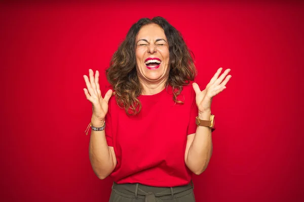 Middle age senior woman with curly hair over red isolated background celebrating mad and crazy for success with arms raised and closed eyes screaming excited. Winner concept