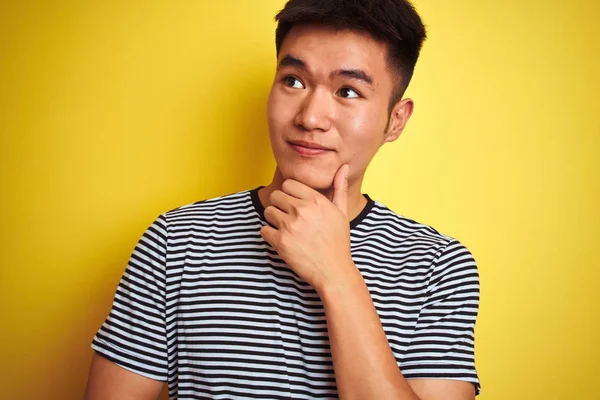Young asian chinese man wearing striped t-shirt standing over isolated yellow background serious face thinking about question, very confused idea