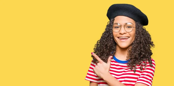 Young beautiful woman with curly hair wearing glasses and fashion beret cheerful with a smile of face pointing with hand and finger up to the side with happy and natural expression on face