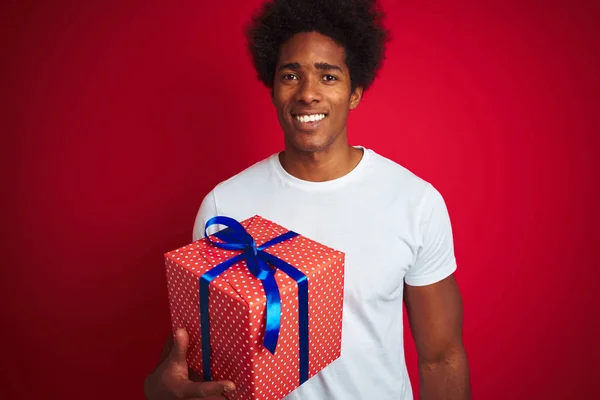 Young african american man holding birthday gift standing over isolated red background with a happy face standing and smiling with a confident smile showing teeth