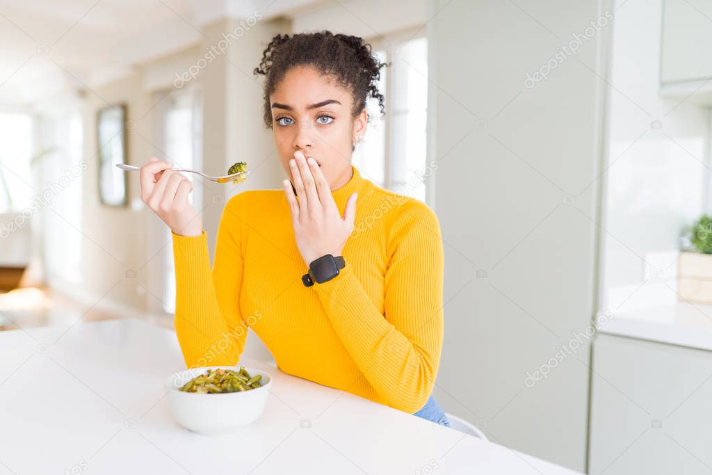 Young african american woman eating healthy green vegatables cover mouth with hand shocked with shame for mistake, expression of fear, scared in silence, secret concept