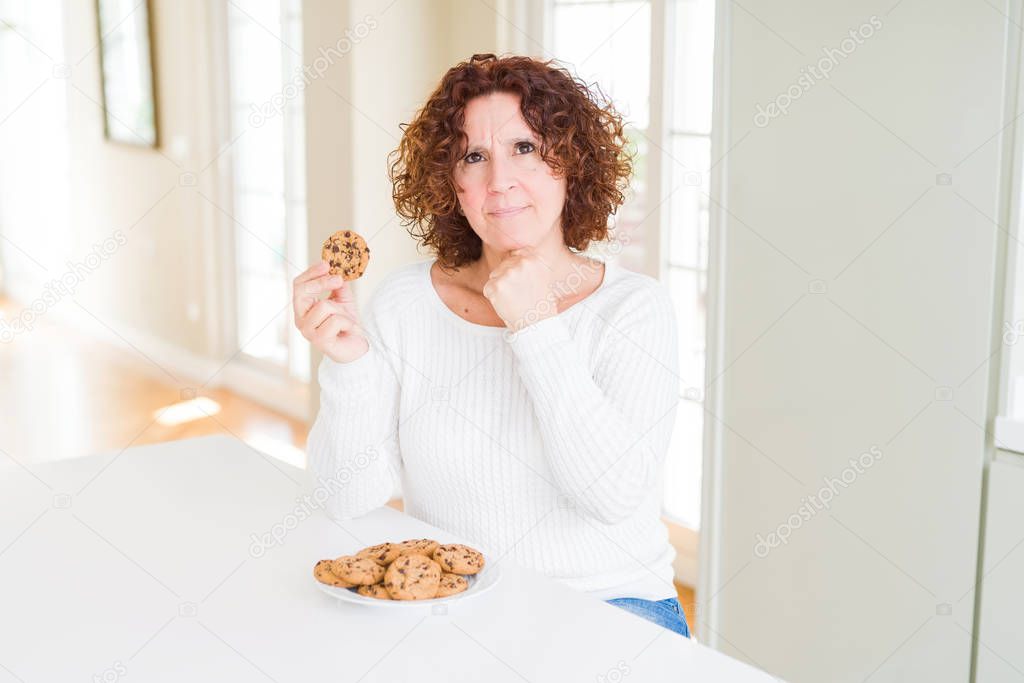 Senior woman eating chocolate chips cookies at home serious face thinking about question, very confused idea