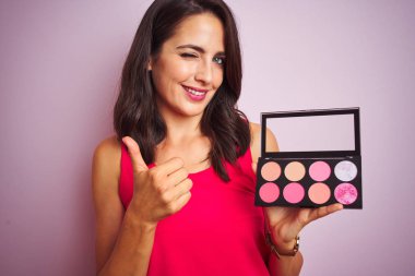 Genç ve güzel bir kadın pembe izole edilmiş arka planda makyaj yapıyor. Gülümsüyor ve gülümsüyor. Tamam işareti yapıyor.