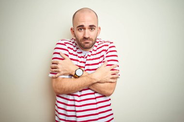 Sakallı genç ve kel bir adam beyaz izole edilmiş arka planda sıradan kırmızı tişörtler giyiyordu. Kış soğuğu yüzünden titriyor ve donuyordu. Yüzünde üzgün ve şok ifadesi vardı.