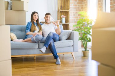 Genç ve güzel bir çift, yeni eve taşındıktan sonra koltukta oturup gülümsüyor ve başparmağıyla yan tarafı işaret ediyor..