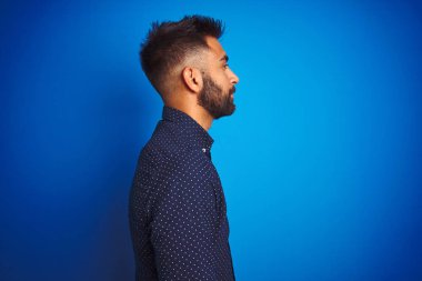 İzole edilmiş mavi arka plan üzerinde duran genç Hintli zarif adam yan bakıyor, rahat poz veriyor kendine güvenen bir gülümsemeyle doğal bir yüze sahip..