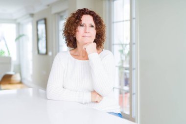 Güzel yaşlı kadın çene üzerinde el ile beyaz kazak giyen soru, dalgın ifade düşünme. Düşünceli bir yüzle gülümsüyor. Şüphe kavramı.