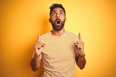 İzole edilmiş sarı arka plan üzerinde duran genç Hintli adam şaşırdı ve şaşırdı. Parmaklarıyla işaret etti ve kollarını kaldırdı..