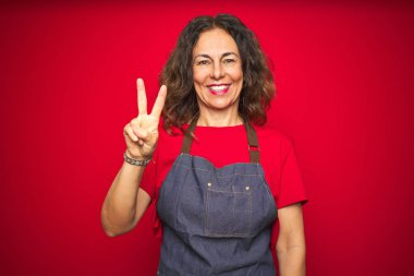 Kırmızı izole edilmiş arka planda önlük giyen orta yaşlı bir kadın iki numaralı parmaklarını gösterip gülümserken kendinden emin ve mutlu görünüyor..