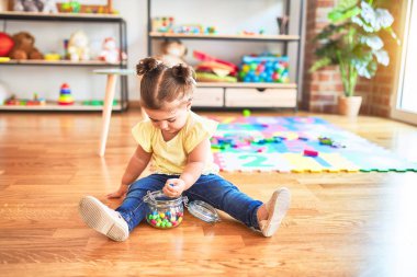 Anaokulunda elinde bir kavanoz çikolata renkli topla yerde oturan güzel çocuk.