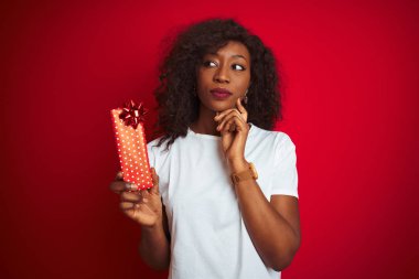 Sevgililer günü hediyesini elinde tutan Afrikalı genç bir Amerikalı kadın izole edilmiş kırmızı arka planda duruyor. Ciddi bir yüz. Soru sormayı düşünüyor.