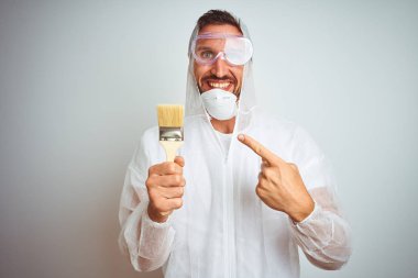 Profesyonel işçi ekipmanı giyen ressam izole edilmiş arka planda fırça tutuyor. Elle ve parmakla göstererek çok mutlu.