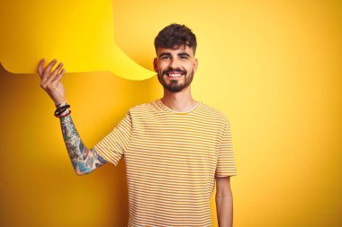 Dövmeli genç adam konuşma balonu tutuyor. Sarı arka planda duruyor. Mutlu bir yüzü var.