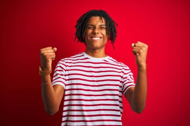 Kırmızı arka plan üzerinde çizgili tişört giyen genç afro bir adam mutlu ve heyecanlı bir şekilde kollarını kaldırıp, gülümseyerek ve başarı için çığlık atarak zafer hareketi yapıyor. Kutlama konsepti.