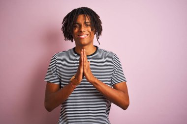 Siyah saçlı, denizci çizgili tişörtlü bir Afro adam izole edilmiş pembe arka planda duruyor ellerini birleştirip af diliyor kendinden emin bir şekilde gülümsüyor..