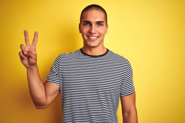 Sarı izole arka plan üzerinde çizgili t-shirt giyen genç yakışıklı adam mutlu yüz zafer işareti yapıyor kamera göz kırpıyor ile gülümsüyor. İki numara.