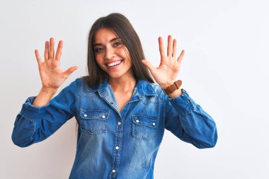 İzole edilmiş beyaz arka plan üzerinde duran sıradan bir kot gömlek giyen genç ve güzel bir kadın on numaralı parmaklarıyla işaret ederken kendinden emin ve mutlu bir şekilde gülümsüyor..