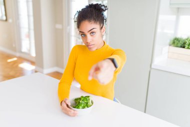 Genç Afro-Amerikalı kız sıcak yeşil brokoli yiyor. Parmağıyla kamerayı işaret ediyor. Ve sen, el işareti, ön taraftan pozitif ve kendinden emin bir hareket.