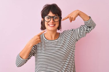 İzole edilmiş pembe arka planda çizgili tişört ve gözlük takan genç ve güzel bir kadın neşeli bir şekilde gülümseyip parmaklarını ve ağzını işaret ediyor. Diş sağlığı konsepti.