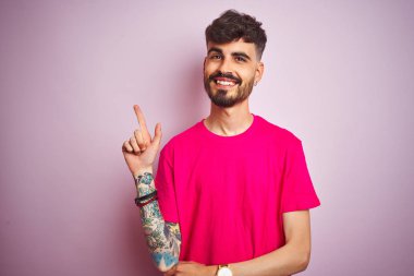 Dövmeli genç adam pembe arka plan üzerinde duruyor yüzünde kocaman bir gülümseme, eli ve parmağı kameraya bakıyor..