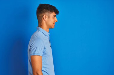 Sıradan polo giyen genç Hintli adam mavi arka planda dikilip yan yana bakıyor, rahat bir görünüm sergiliyor, kendine güvenen bir gülümsemesi var..