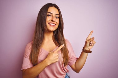 İzole edilmiş pembe arka planda duran tişört giyen genç ve güzel bir kadın gülümseyip kameraya bakıyor ve iki eliyle ve parmaklarıyla kenarı işaret ediyor..