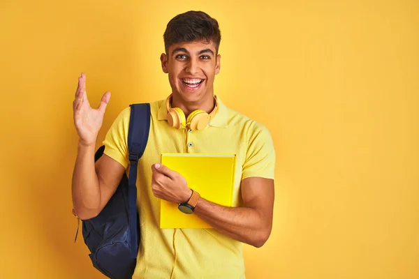 孤立した黄色の背景にバックパックのヘッドフォンノートを身に着けているインドの学生の男は非常に幸せと興奮 大きな笑顔で叫んで勝利を祝う勝者式と手を上げ — ストック写真