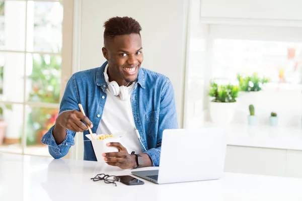 Handsome young african business man eating delivery asian food and working using computer, enjoying noodles smiling
