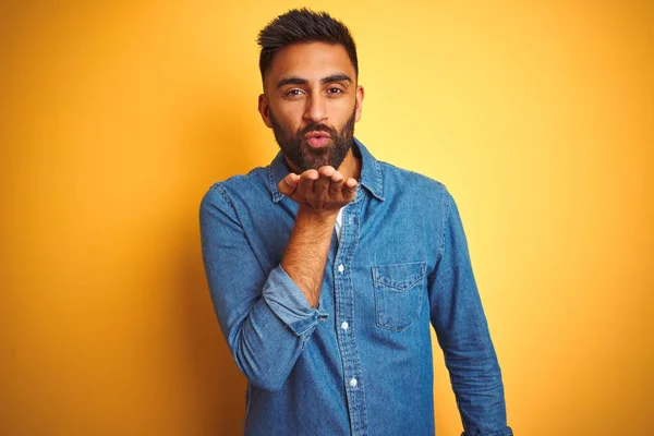 Young indian man wearing denim shirt standing over isolated yellow background looking at the camera blowing a kiss with hand on air being lovely and sexy. Love expression.