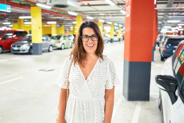 Yeraltı otoparkında araba ve ışıkların etrafında kendine güvenen genç bir kadın.