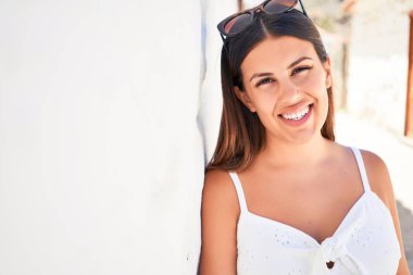 Güzel kız beyaz duvara yaslanıyor, genç arkadaş canlısı kadın mutlu bir yaz gününde gülümsüyor.