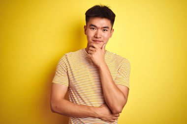 İzole edilmiş sarı arka planda duran tişört giyen Asyalı genç adam kameraya kollarını kavuşturup gülümseyerek ve çenesini kaldırarak bakıyor. Olumlu düşünüyorum..