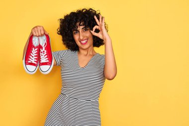 Kıvırcık saçlı genç Arap kadın izole edilmiş sarı arka planda spor ayakkabı tutuyor. Tamam işareti yapıyor, mükemmel bir sembol.