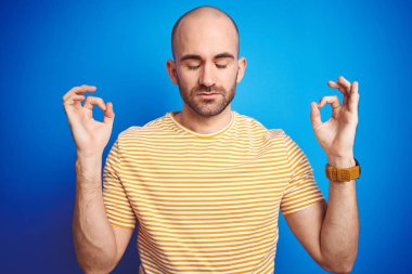 Mavi yalıtılmış arka plan üzerine günlük tişört giyen sakallı genç kel adam rahatla ve parmaklarıyla meditasyon hareketi yaparken gözleri kapalı gülümse. Yoga konsepti.