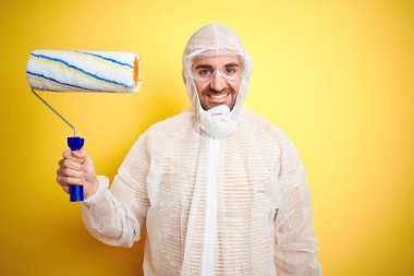 Boyacı kıyafeti giyen genç bir adam ve izole edilmiş sarı arka planda elinde boya rulosu tutan mutlu bir yüzü olan ve kendine güvenen bir gülümsemeyle dişlerini gösteren bir adam.
