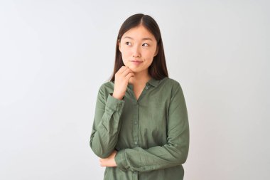 Sıradan yeşil tişört giyen genç Çinli bir kadın izole edilmiş beyaz arka planda duruyor ve elleri çenesinde soru sormayı düşünüyor, düşünceli bir ifade. Düşünceli bir yüzle gülümsüyor. Şüphe kavramı.