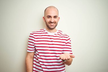 Sakallı genç ve kel bir adam, beyaz izole edilmiş arka planda günlük kırmızı tişört giyiyor. Gülümsüyor, mutlu bir avuç içi dolusu yardım ve kabullenme sunuyor..