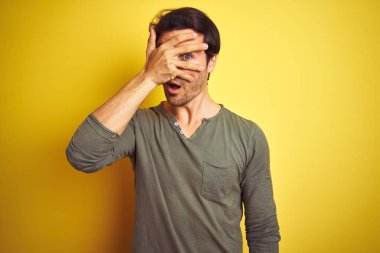 Genç ve yakışıklı bir adam günlük tişört giyip izole edilmiş sarı arka planda dikiliyor. Şok olmuş bir şekilde yüzünü ve gözlerini kapıyor. Utanmış bir ifadeyle parmaklarının arasından bakıyor..