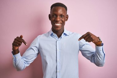 Mavi zarif gömlekli Afro-Amerikan bir adam pembe arka planda duruyor. Yüzünde bir gülümsemeyle, gururlu ve mutlu bir şekilde kendini işaret ediyor..