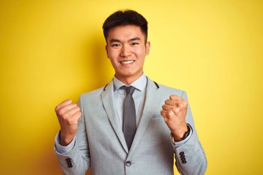 Asyalı Çinli işadamı takım elbise ve kravat takıp izole edilmiş sarı arka planda bekleyerek kollarını kaldırıp, başarıyı kutluyor ve şaşırıyor. Kazanan konsept.