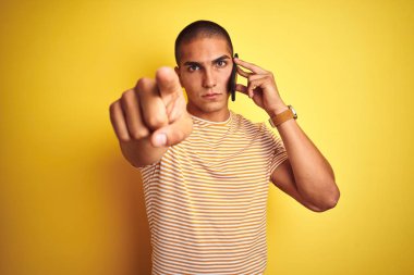 Genç yakışıklı adam, sarı arka plan üzerinde akıllı telefon kullanarak konuşuyor. Kamerayı işaret ediyor ve sen, el işareti, ön taraftan pozitif ve kendinden emin bir hareket.