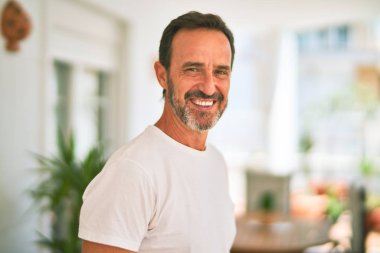 Orta yaşlı yakışıklı bir adam terasta dikilmiş gülümseyerek günlük tişört giyiyor.