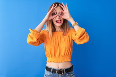 Güzel kızıl saçlı kadın turuncu günlük tişört giyiyor mavi arka planda dikiliyor dürbün gibi güzel hareketler yapıyor, gözler parmaklarından bakıyor. Çılgın ifade.