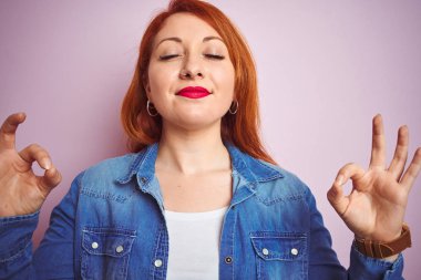 Youg güzel kızıl saçlı kadın izole pembe arka plan üzerinde duran denim gömlek giyen rahatlayın ve gözleri parmakları ile meditasyon jest yapıyor kapalı gülümseyerek. Yoga konsepti.