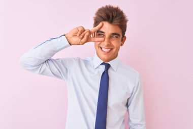 Genç yakışıklı işadamı gömlek ve kravat takıyor pembe arka planda dikiliyor barış sembolü yapıyor parmakları yüzünde, neşeyle gülümsüyor zaferi gösteriyor