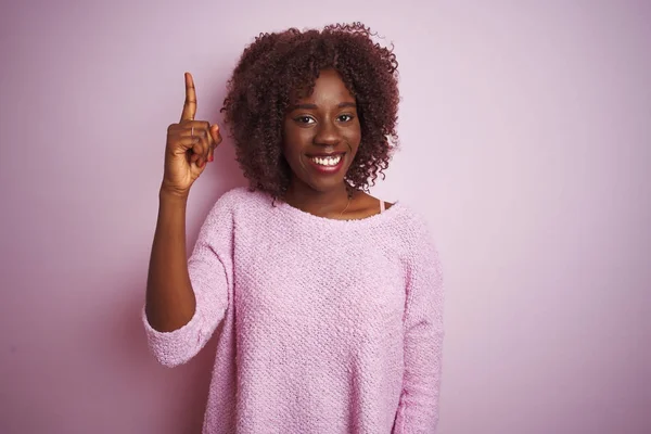 Young african afro woman wearing sweater standing over isolated pink background pointing finger up with successful idea. Exited and happy. Number one.
