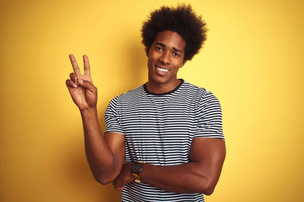 African american man with afro hair wearing navy striped t-shirt over isolated yellow background smiling with happy face winking at the camera doing victory sign. Number two.