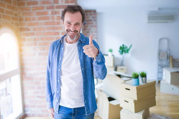 Middle age senior man moving to a new house packing cardboard boxes doing happy thumbs up gesture with hand. Approving expression looking at the camera with showing success.