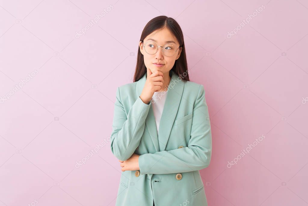 Young chinese businesswoman wearing jacket and glasses over isolated pink background with hand on chin thinking about question, pensive expression. Smiling with thoughtful face. Doubt concept.